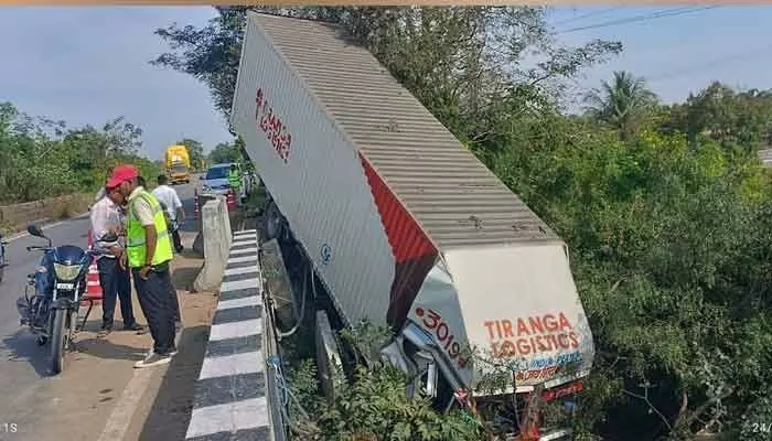 A speeding container lorry on NH44 lost control, crashed through barriers, and fell into Haldi Vagu. Driver and cleaner escaped with minor injuries.